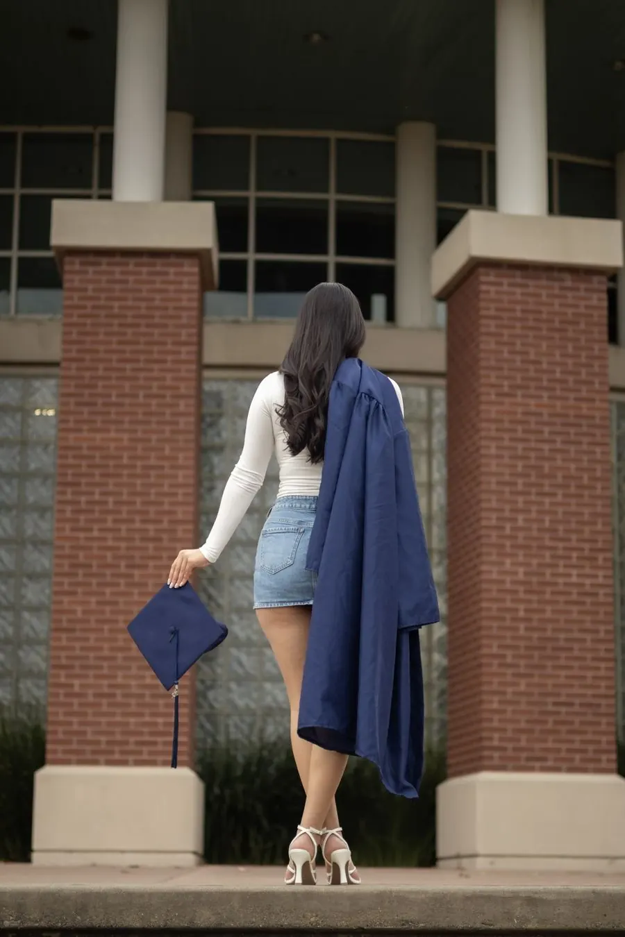 Graduate seen from behind holding her cap, gown over her shoulder, in front of brick columns
