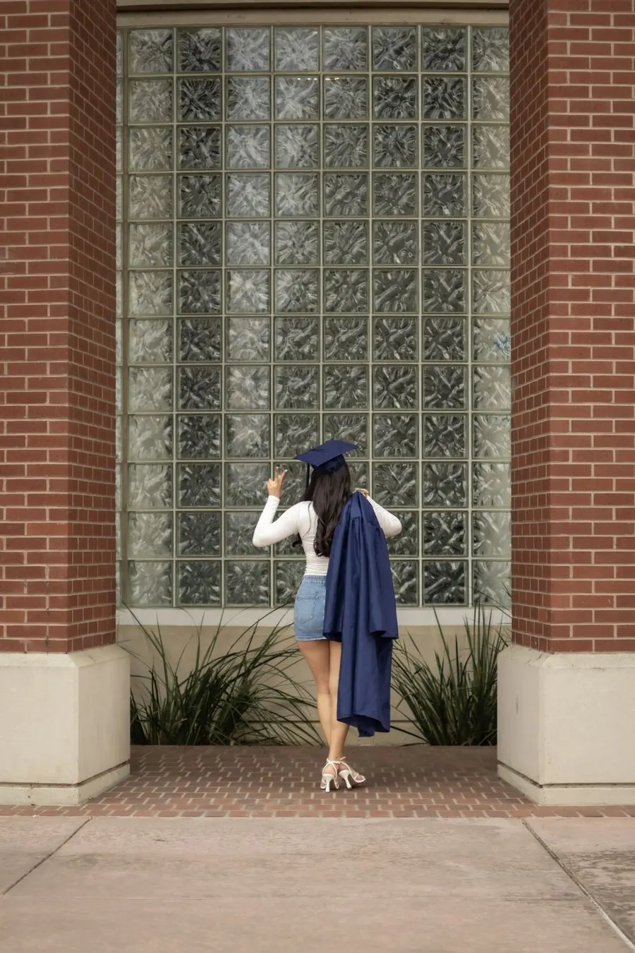 Graduate seen from behind, gown over her shoulder, flashing a peace sign at a glass block window