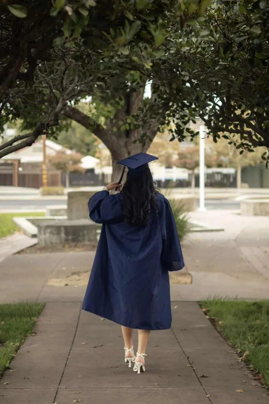 Graduate in cap and gown walking away down a sidewalk under trees