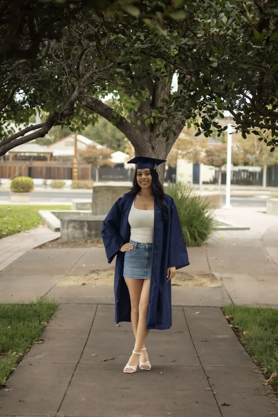 Graduate in cap and gown smiling on a tree-lined sidewalk